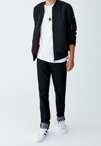 Blouson aviateur noir avec fermeture éclair et poches latérales, t-shirt blanc, pantalon en jean foncé, et baskets blanches avec des accents noirs.