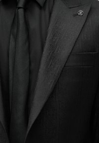 Μαύρο σακάκι με διακριτικό μοτίβο, μονόχρωμο μαύρο πουκάμισο, μαύρη γραβάτα με υφή και ασημένια στρογγυλή καρφίτσα στο πέτο.