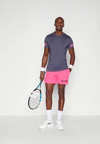 Muž v purpurové košili, růžových šortkách, bílých ponožkách a botách drží tenisovou raketu a stojí před hladkým šedým pozadím.