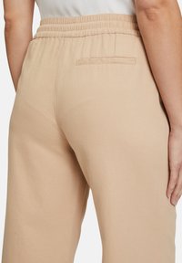 Beige, hooggeplaatste broek met een elastische tailleband en een enkele achterzak. Gladde textuur met een ontspannen pasvorm.
