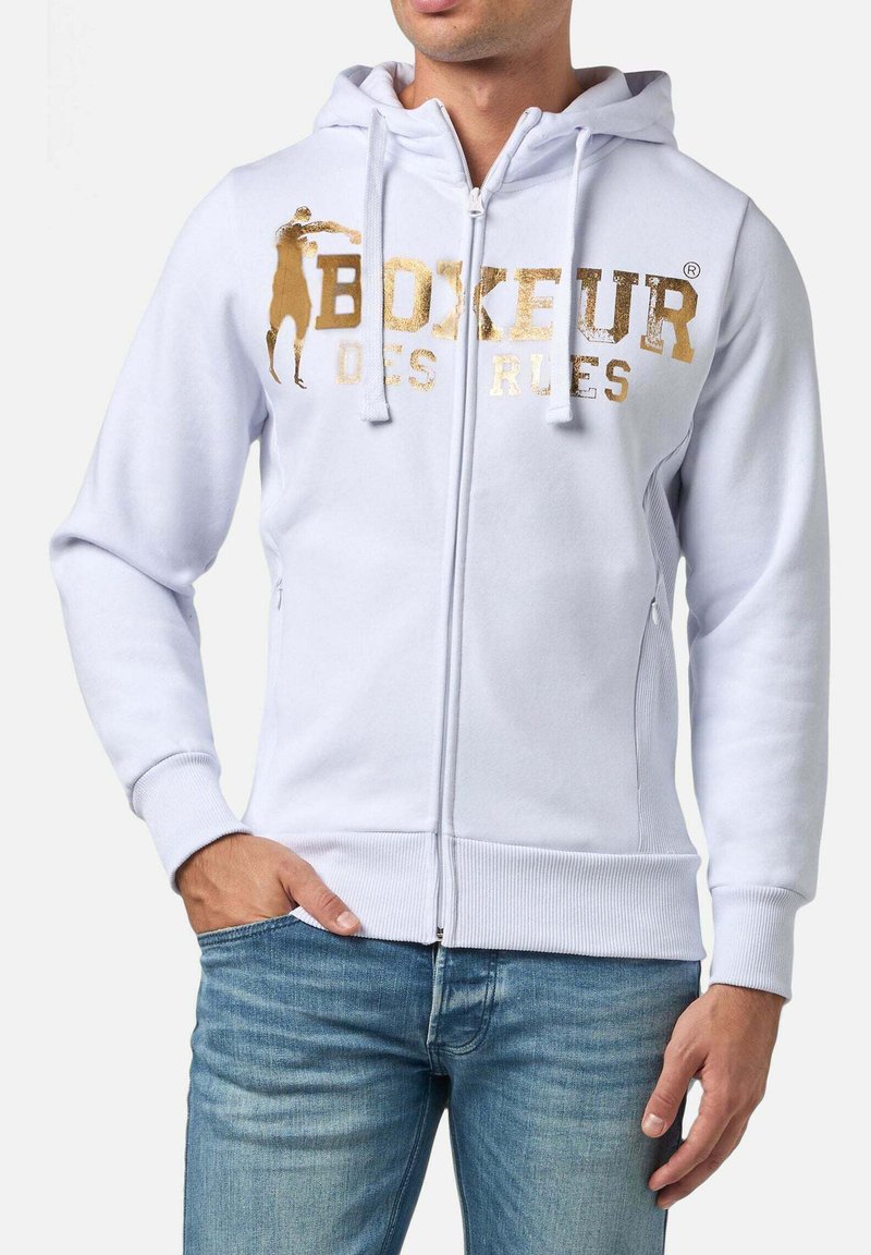 Felpa bianca con zip e logo dorato stampato "BOXEUR DES RUES" e una grafica. Presenta un cappuccio con cordino e polsini a costine. Indossata con jeans chiari.