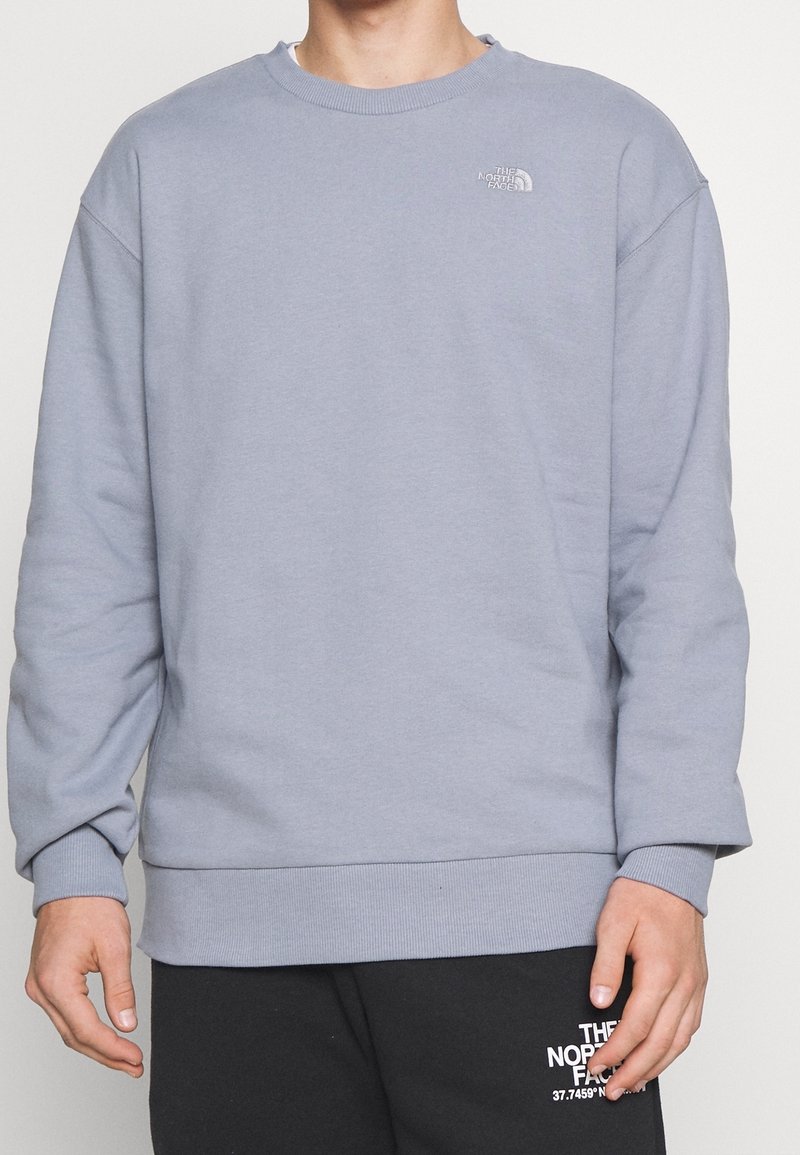 Homme portant un sweat-shirt gris clair col rond et un pantalon de jogging noir, tous deux avec le logo blanc The North Face sur la poitrine et la jambe.