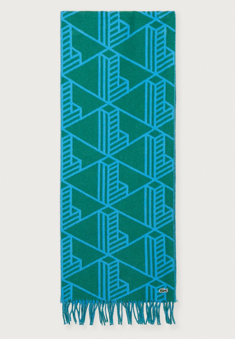 Bufanda verde azulado con un patrón geométrico azul que presenta triángulos y rayas, terminada con bordes con flecos y una pequeña etiqueta de logo.