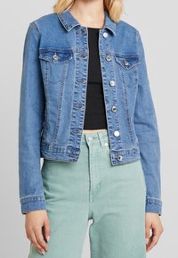 Jeansjacka i ljusblått, med klassisk krage, knappstängning, två bröstfickor och sydda detaljer. Bärs över en svart topp.