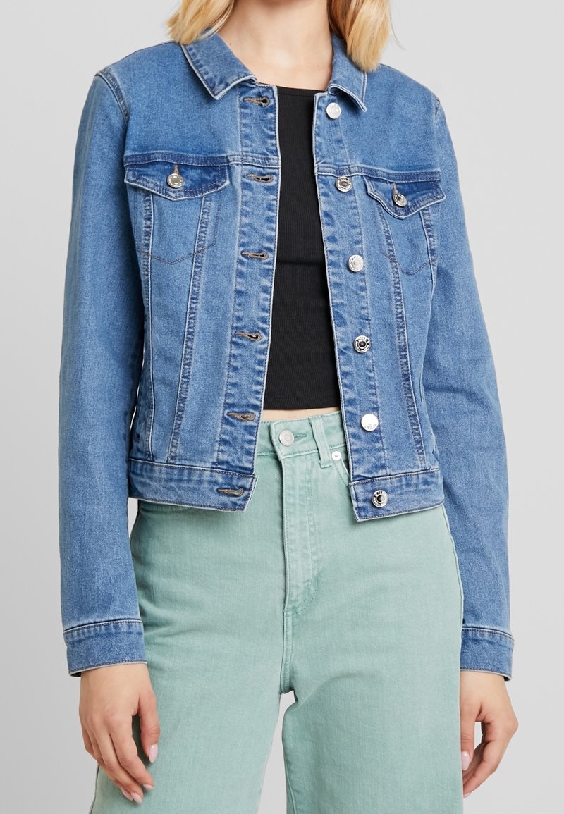 Jeansjacka i ljusblått, med klassisk krage, knappstängning, två bröstfickor och sydda detaljer. Bärs över en svart topp.