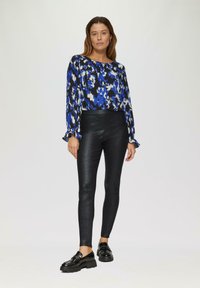 Blouse à motifs bleus et noirs avec des manches volantes, assortie à des leggings noirs brillants et des mocassins noirs épais. Fond simple.