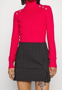 Femme portant un pull à col roulé rose vif avec des boutons aux épaules et une mini-jupe noire à rayures fines avec une petite fente à l'avant.