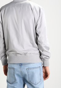 Jasnoszary bomber jacket z ryflowanym kołnierzem i mankietami, wykonany z gładkiego, błyszczącego materiału. W zestawie z jasnoniebieskimi dżinsami.
