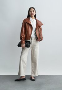 Νεαρή γυναίκα στέκεται, φορώντας καφέ δερμάτινο μπουφάν, λευκό πουκάμισο, εκρού παντελόνι με φαρδιά μπατζάκια, μαύρα φλατ παπούτσια και κρατάει μια μαύρη τσάντα φάκελο με καπιτονέ σχέδιο.