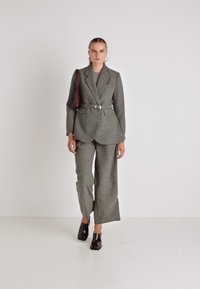 Sivi tvid jakna s remenom, usklađene široke hlače, u paru s tamnocrvenom torbicom. Prikazano na jednostavnoj bijeloj pozadini.