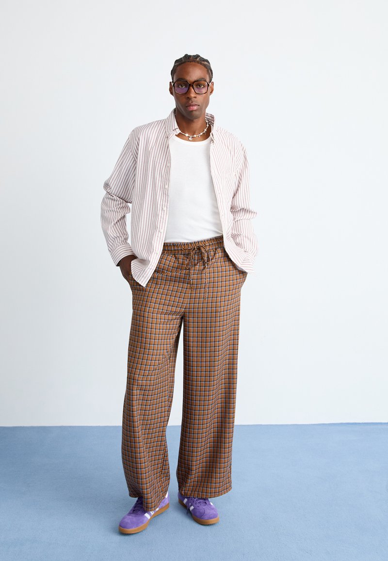 Gestreiftes, leichtes Hemd, weißes Tanktop, karierte breite Hose in Braun und Orange sowie lila Schuhe. Minimalistische Accessoires und Brille.