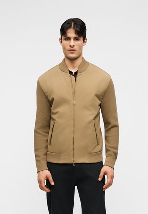 Jonge man met kort donker haar draagt een beige bomberjack met gebreide mouwen en zwarte broek, staand tegen een effen lichte achtergrond.