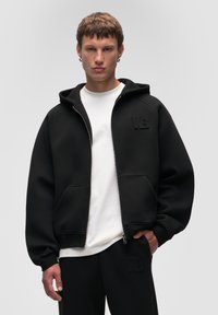 Felpa nera con zip, grande cappuccio, tasche anteriori e logo "WB" in rilievo. Indossata sopra una maglietta bianca e abbinata a pantaloni neri.