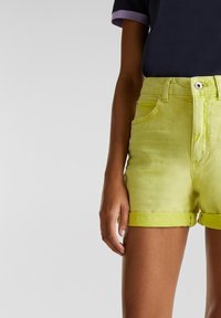 Gros plan d'une personne portant un short en jean jaune vif à ourlet roulé et une chemise sombre à manches courtes avec un bord violet sur les manches.