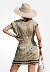 Häkelüberwurf für den Strand in Beige mit schwarzem Saum, mit lockerem Schnitt, kurzen Ärmeln und gewelltem Saum. Kombiniert mit einem Strohhut und einer Tasche.