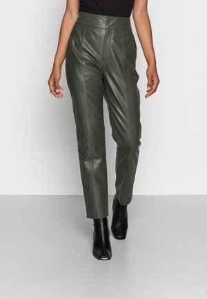 Femme portant un pantalon en cuir vert olive taille haute et des bottines noires, debout devant un fond clair uni.