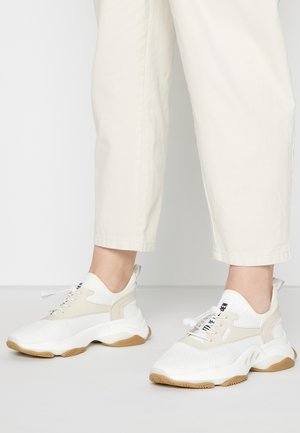 Witte en beige chunky sneakers met rubberen zolen, gedragen met licht crèmekleurige cropped broeken op een effen lichte achtergrond.