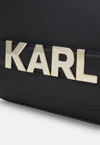 Borsa nera in pelle con un vistoso logo "KARL" in tono oro sulla parte frontale, texture liscia con dettagli di cucitura.