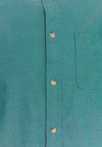 Turquoise overhemd met zeven knopen, een borstzak en een getextureerde stof met een geweven patroon.