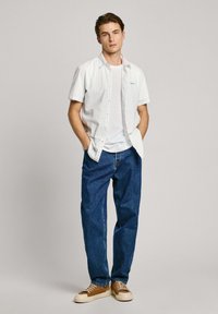 Camisa blanca de manga corta con rayas azul claro, llevada sobre una camiseta blanca. Combinada con unos vaqueros azules y zapatillas marrones.