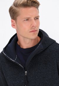 Fleece jaciņa ar rāvējslēdzēju tumši zilā krāsā, ar strukturētu audumu un kontrastējošu iekšējo oderi. Iespējas: augsts apkakle un redzams rāvējslēdzējs.