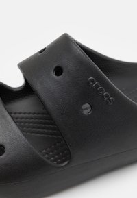 Črne Crocs sandale imajo teksturirano, fleksibilno zasnovo z zračnimi odprtinami, gladko notranjo oblogo in embosiran logotip na trak.