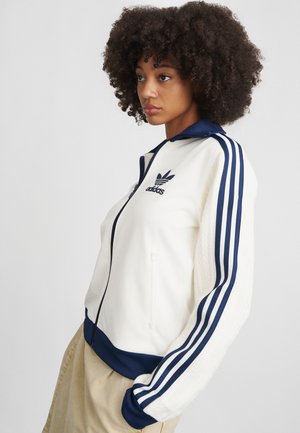 Casaco branco com fecho zip e detalhes em azul marinho, com três riscas nas mangas. Apresenta um logo bordado no peito e punhos canelados.