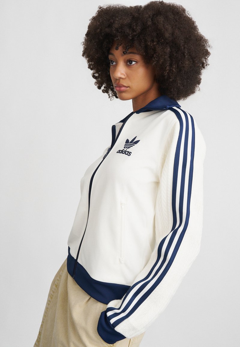 White zip-up jacket with navy blue accents and three stripes on sleeves. Features an embroidered logo on the chest and ribbed cuffs.