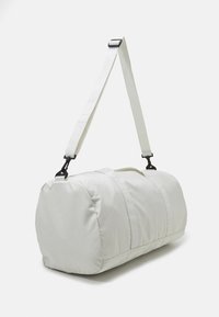 Biały worek podróżny wykonany z miękkiego materiału, o okrągłym kształcie, z regulowanym paskiem i dwoma uchwytami do noszenia. Gładka tekstura z minimalistycznym wzornictwem.