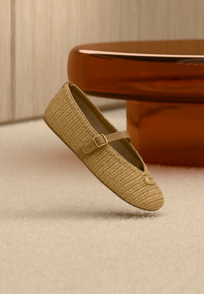 Scarpa ballerina beige intrecciata modello Mary Jane con cinturino in pelle e fibbia, posizionata sopra una moquette chiara, mobili arrotondati marroni sullo sfondo.