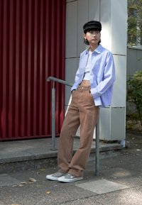 Chemise rayée bleu clair et blanche, crop top blanc, pantalon large marron, baskets grises et casquette noire. Se tenant près d'une balustrade en métal.