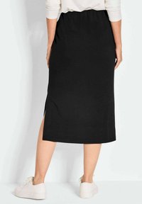 Jupe midi noire avec une fente latérale, fabriquée en tissu doux. Elle présente une taille élastique et une texture lisse, portée avec des baskets blanches.