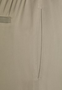 Light beige fabric with a smooth texture, featuring a side pocket and subtle stitching details. The design includes slight pleating at the waist.