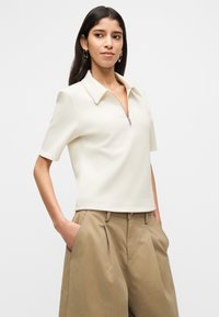 Γυναίκα με μακριά σκούρα μαλλιά, ντυμένη με εκρού μπλούζα με φερμουάρ και κοντά μανίκια, και χαλαρά μπεζ παντελόνια, με τα χέρια στις τσέπες, στέκεται μπροστά σε απλό φόντο.