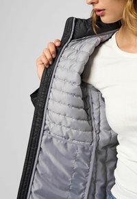 Černá vnější bunda se šedým prošívaným vnitřkem, ukazující hladkou texturu a kontrastní barvy. Bunda je rozepnutá na zip, odhalující detaily.