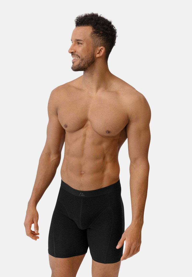 Shirtless man with short curly hair smiles looking to the side, wearing fitted black boxer briefs against a plain background.