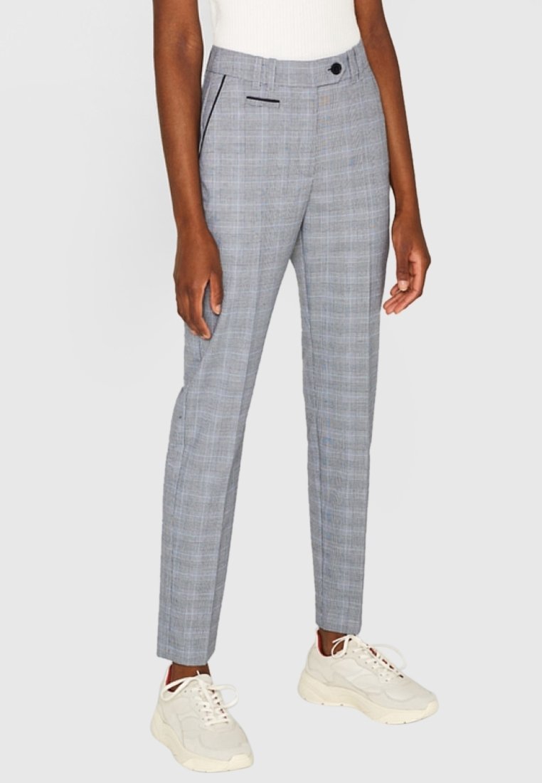 Femme portant un pantalon slim à carreaux gris, des baskets blanches et un haut sans manches blanc sur un fond uni.