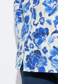 Kukkamainen paita, jossa on sinisiä ja vaaleansinisiä kuvioita kermaisen taustan päällä; siinä on sivureikä ja teksturoitu kangas. Tummat farkut näkyvät alapuolella.