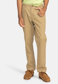 Pantalons en coton beige avec une coupe droite, dotés d'un bouton de fermeture, de passants de ceinture et de poches latérales, associés à des chaussures en daim marron.