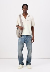 Light beige short-sleeve shirt with black accents, loose blue denim jeans, and cream-colored sneakers. Model carries a gray crossbody bag.