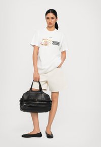 Tricou alb cu grafică de ceașcă și textul "SHIRT", asortat cu pantaloni scurți crem. Geantă de mână din piele neagră, cu un design structurat și bretele.