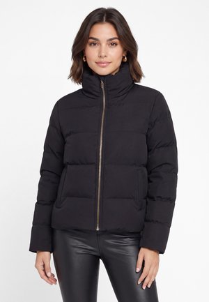 Giovane donna con capelli scuri fino alle spalle indossa una giacca imbottita nera con zip frontale e pantaloni neri in ecopelle.