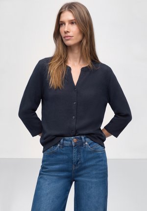 Femme debout, les mains derrière le dos, portant un chemisier noir boutonné à manches trois-quarts et un jean bleu taille haute, sur fond blanc.