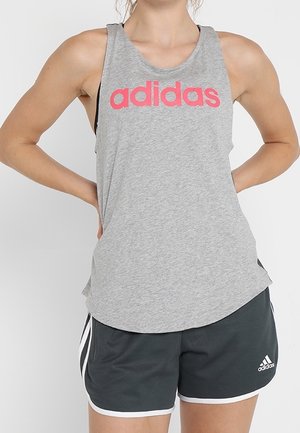 Osoba nosící šedé tílko Adidas s růžovým logem a černé šortky Adidas s bílým lemem, ruce opřené o boky.