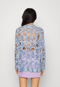 Lettstrikket sweater i blå gradient med udskårne blomster mønstre. Har lange ærmer og en løs pasform, lagd oven på en lilla kjole.