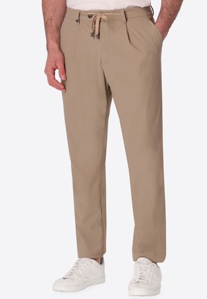 Pantaloni beige a gamba stretta con vita con bottone e coulisse, tasche laterali, indossati con scarpe da ginnastica bianche e camicia bianca infilata.