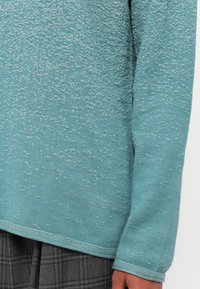 Ein türkisfarbener Pullover mit einer strukturierten Oberfläche, die kleine silberne Punkte aufweist. Das Design hat einen geraden Schnitt und einen gerippten Saum.