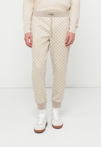 Φόρμα με μπεζ βάση που διαθέτει επαναλαμβανόμενο μοτίβο λογότυπου, ελαστική μέση με κορδόνι και ριμπ μανσέτες. Συνδυάζεται με λευκά αθλητικά παπούτσια.