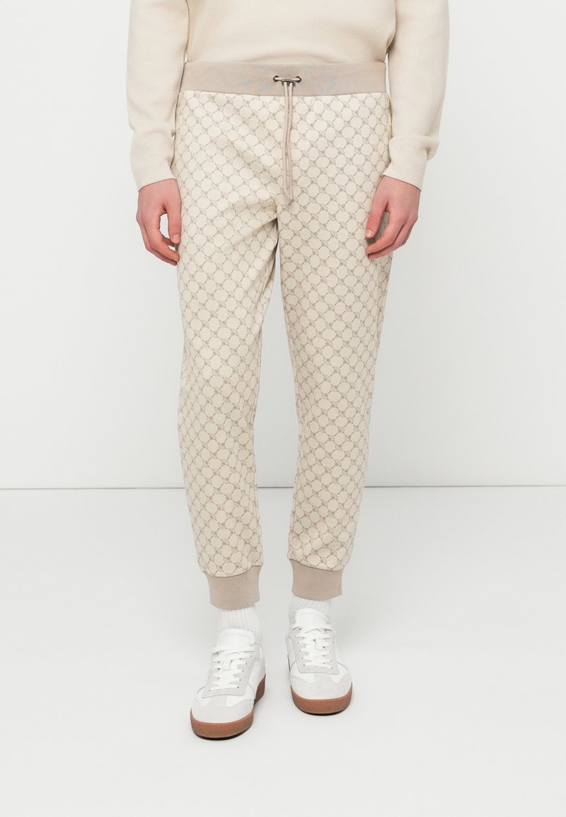 Φόρμα με μπεζ βάση που διαθέτει επαναλαμβανόμενο μοτίβο λογότυπου, ελαστική μέση με κορδόνι και ριμπ μανσέτες. Συνδυάζεται με λευκά αθλητικά παπούτσια.