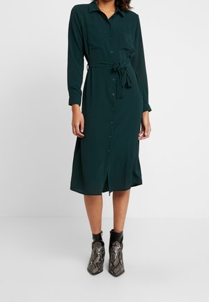 Woman wearing a dark green, long-sleeve, button-up midi dress with a tied waist belt and patterned ankle boots.